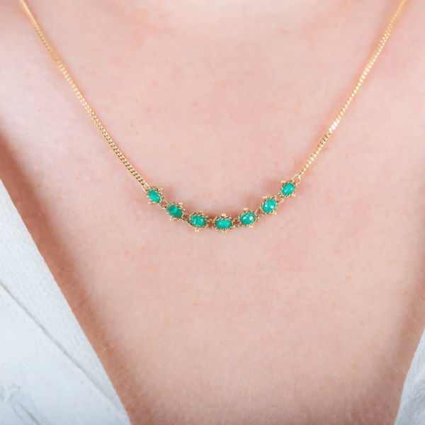 18k yellow gold chain necklace featuring a delicate row of seven handwoven emerald gemstones worn on a neck.