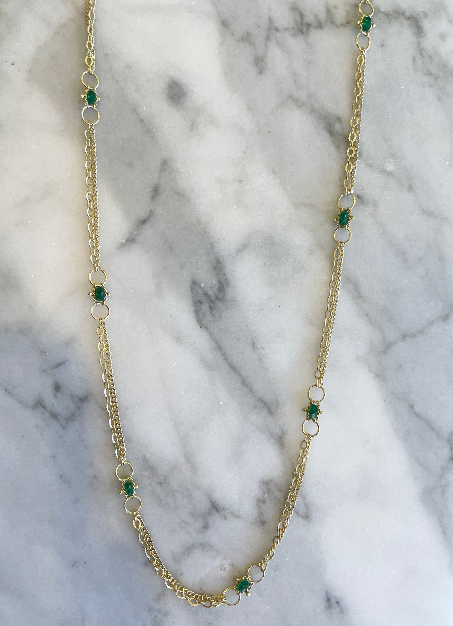 18k yellow gold chain necklace featuring hand-woven emerald stones on a marble background.