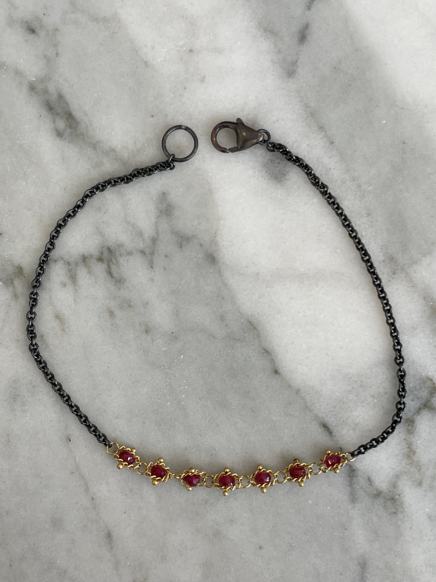 Oxidized sterling silver chain bracelet with seven faceted ruby beads woven into 18k yellow gold on a marble surface.