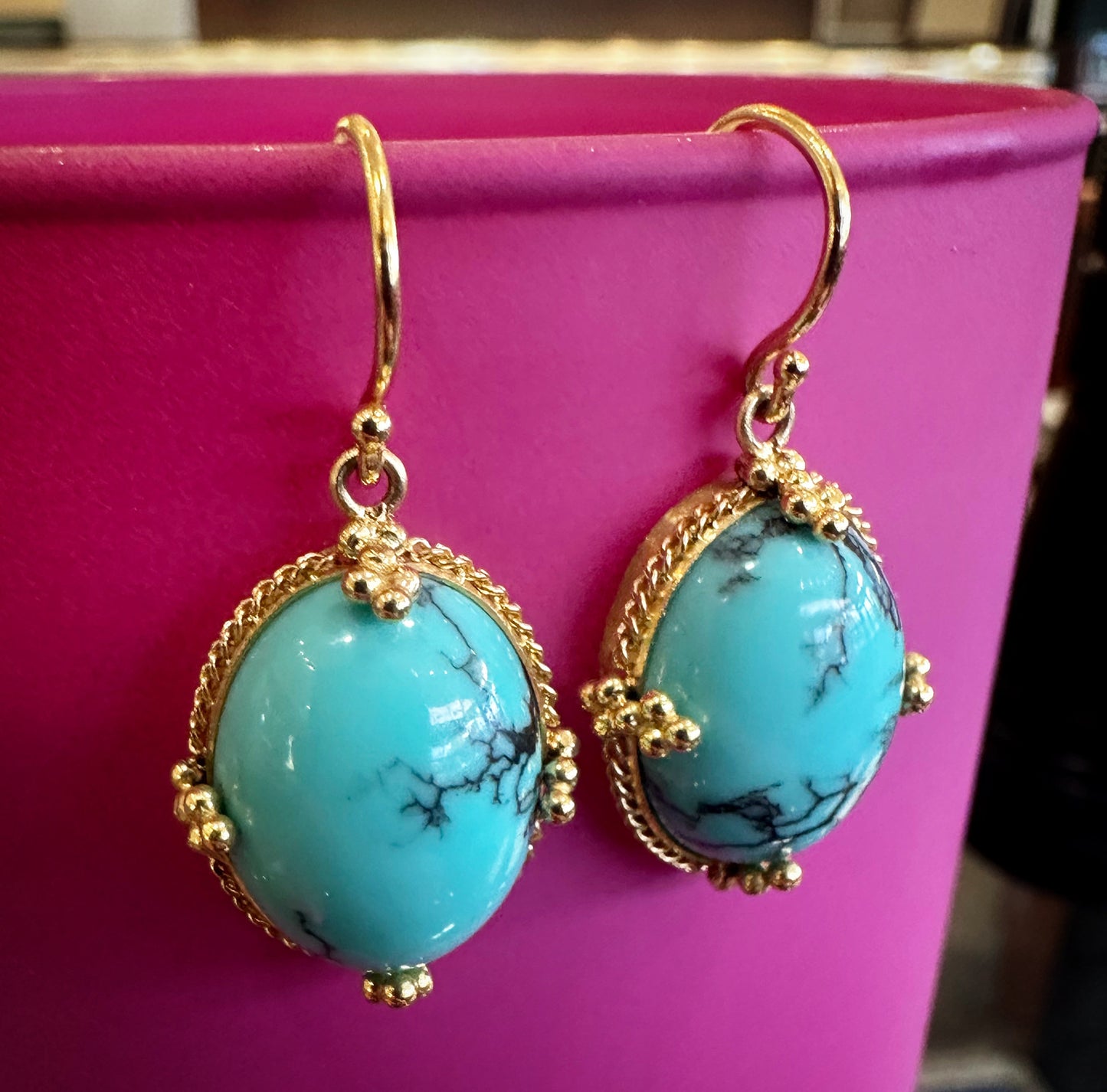 Oval turquoise earrings with black veining set in ornate 18k yellow gold frames on a bright pink background.