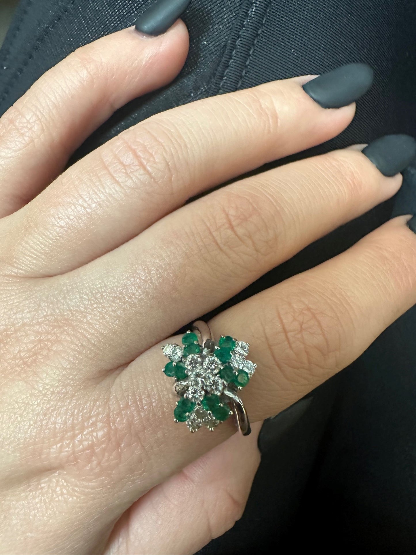 14k white gold cluster ring featuring a round emerald surrounded by 13 brilliant-cut diamonds on a finger.